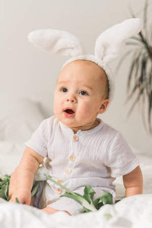 Cute funny baby with bunny ears, with a tulips at home on a bed of white bedding. Easter greeting...
