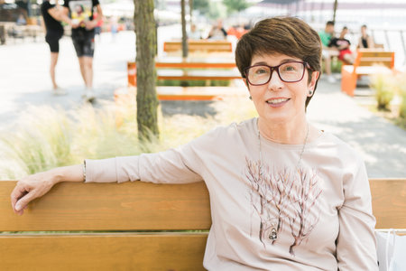 Outdoor portrait of beautiful and elegant middle age 55 - 60 year old woman wearing long sleeve top sitting on bench on the street. Copy space and empty place for text advertisingの写真素材