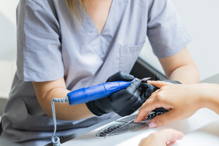 Manicurist uses an electric drill to remove old gel polish from nails. A woman is getting a manicure of nails. The beautician files the clients nails. professional manicure tool. Close-upの写真素材