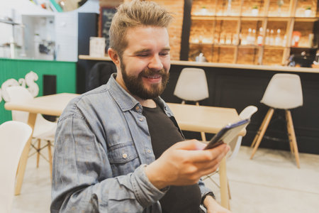 Bearded male using smartphone at modern coffee shop and chatting online messaging on mobile phone. Social networks and reading blog conceptの写真素材