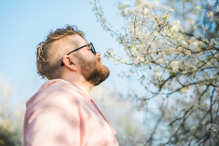 Portrait of curly millennial man inhales the fragrance of spring flowers of blooming jasmine or cherry tree. Spring time concept. Copy spaceの写真素材