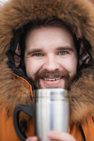 Close-up portrait handsome bearded millennial man in winter clothes and with thermos snow outdoor. Cold season and hot beverage in winter timeの写真素材