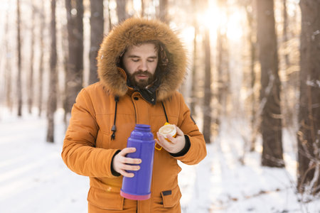 Man pours hot tea from a thermos into a cup walking in snowy frozen winter forest at sunset....