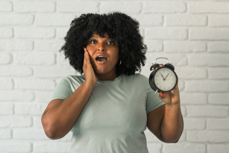 Surprised african american woman holding alarm clock amazed with open mouth and surprise happy face...