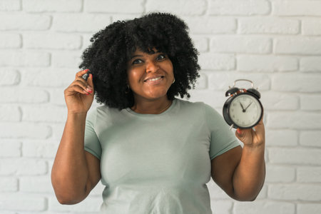 Happy african american woman holding alarm clock amazed and surprise happy face at home, on brick...