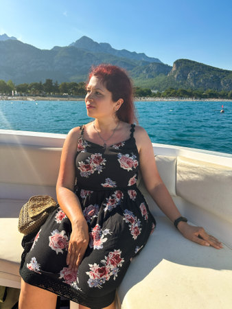 Woman in a black dress sits on a boat under the bright sun, with a scenic view of mountains and the...