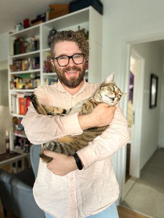 Warm moment between a millennial man and his cat, bond and affection shared in a cozy home environment. The background features a bookshelf at home. Pet and friendship conceptの写真素材