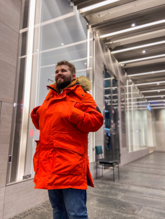 Millennial man in bright orange parka stands confidently on a city street, framed by architectural arches and urban atmosphere in cold season. Modern fashion and classic city charm, emphasizing individuality and styleの写真素材