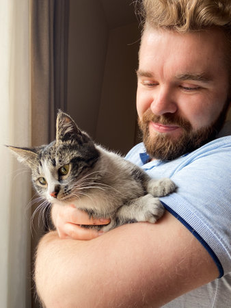 Millennial man gently holds a cat by a sunlit window, capturing a moment of serene companionship....