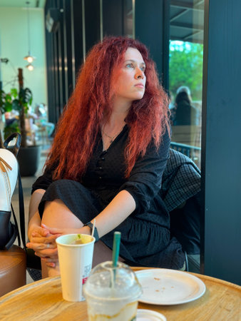 Millennial woman with red hair sits thoughtfully by a large window, gazing outside. Cozy cafe...