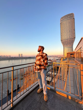 Man stands thoughtfully on a balcony, overlooking a river with a modern skyscraper in Belgrade city during a serene sunset. the warm glow of the evening light casts a peaceful ambiance over the urban landscapeの写真素材
