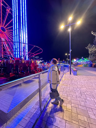 Millennial woman in a fluffy jacket stands illuminated by the colorful lights of a carnival ride in amusement park, gazing upwards with a joyful expression. Winter festive atmosphereの写真素材