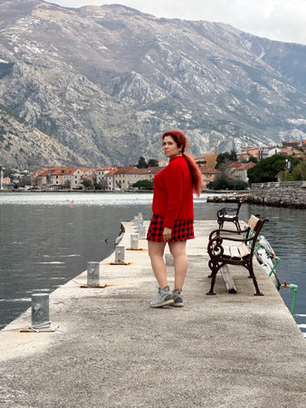 Millennial woman in red standing on a pier against a backdrop of majestic mountains and cloudy skies. a serene and contemplative moment captured by the waterfront.の写真素材