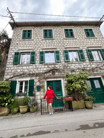 Charming stone building with green shutters and millennial woman in red standing in front. a...