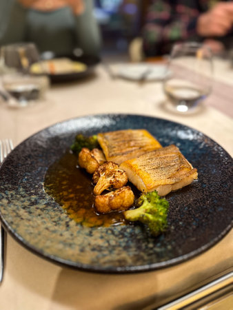 Plated gourmet dish features succulent fish fillets accompanied by roasted cauliflower and broccoli,...