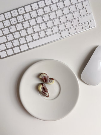 Top view white workspace with keyboard and plate of chocolates. The clean, minimal design emphasizes...