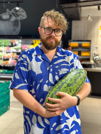 Millennial man holding a watermelon in a grocery store. Moment of everyday life with a vibrant display of produce and personal styleの写真素材