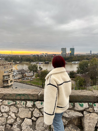 Woman in a cozy jacket overlooks a serene river and urban skyline at sunset in autumn season. Scene captures a moment of tranquility and reflection amidst an expansive cityscapeの写真素材