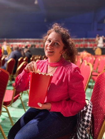 Cheerful woman in a pink blouse enjoys popcorn while seated in a vibrant theater setting. Red chairs...