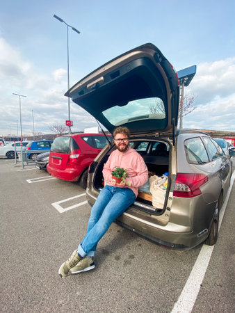 Millennial man in a pink hoodie sits casually on the edge of an open car trunk in a parking lot....