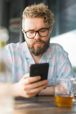 Millennial man with glasses and a patterned shirt sits at an outdoor cafe, focused on his smartphone. Technology and Relaxed summer moments conceptの写真素材