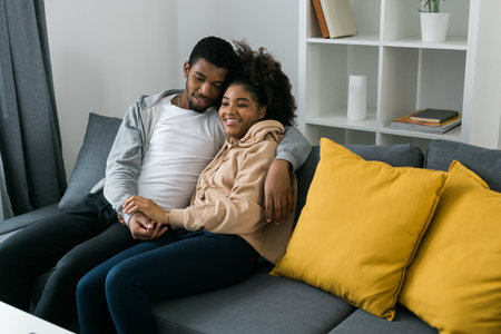 African American couple sits on a cozy gray sofa, embracing each other and enjoying a peaceful moment at home. Love and intimacy and a sense of comfort in a modern and inviting interiorの写真素材