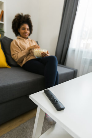 African american woman sits on a gray sofa, enjoying a bowl of popcorn while watching a movie or...