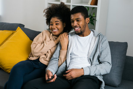African American couple relaxes on a cozy sofa, sharing a joyful moment while watching a show with a...