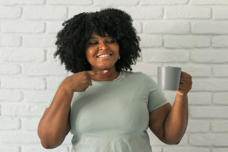 African American cheerful woman holding a gray mug and point on it, showcasing positivity and...