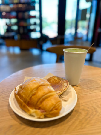 Cozy breakfast scene with a croissant, pancake and matcha latte served on a wooden table. Blurred background of a cafe adds depth and evokes a sense of warmth and relaxation in a casual dining setting. Coffee shop conceptの写真素材