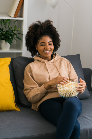 Young African American woman holds a bowl of popcorn while enjoying a television show. Bright smile radiates happiness and relaxation in a cozy home. TV, series and streaming service conceptの写真素材