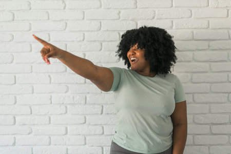 Portrait of a happy young African American woman pointing finger away at copy space over brick wall backgroundの写真素材