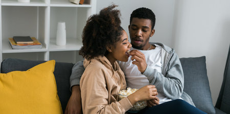 African American couple shares a playful and relaxed moment on the couch, with the man feeding popcorn to the woman. TV, series and streaming service conceptの写真素材