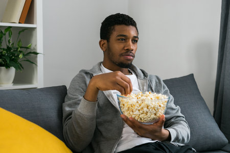 African American man sits alone on the couch and enjoying popcorn while watching television and streaming serviceの写真素材
