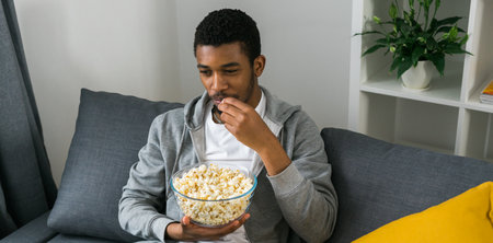 Banner African american man focuses on the television while eating popcorn, fully immersed in show....