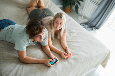 Young couple in love playing video games while lying on the bed in the bedroom, having fun and enjoying each other on their weekends. Technologies and gaming conceptの写真素材