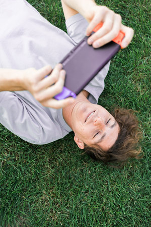 Young man smiling confident playing video game at park lying on a grass. Technology and gaming...