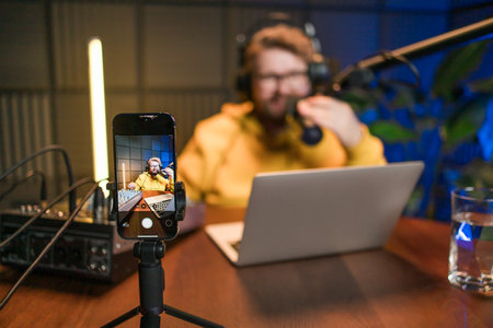 Live streaming with smiling millennial man as radio presenter with microphone, talk show or entertainment. Male person content creator and happy on podcast host in studio for recording audio or social mediaの写真素材