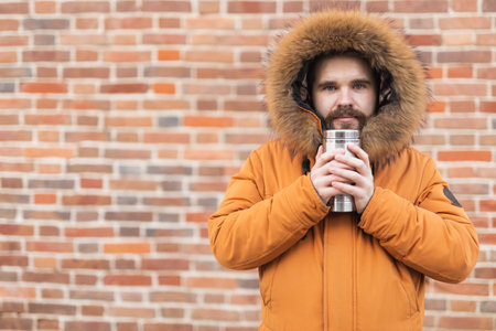 Man with cup looks away on winter street near brick wall with copy space. Seasonal comfort, morning routine, and emotional reflection outdoors.の写真素材