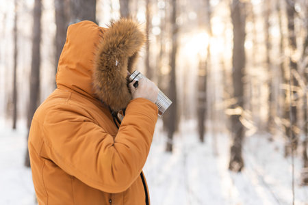 Man drinks from thermos in snowy winter forest during sunrise. Solitude, warmth and peaceful outdoor routine in a winter landscape. Copy spaceの写真素材