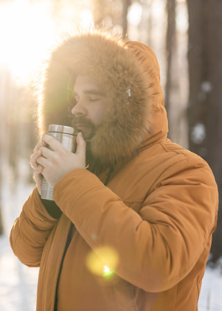 Man warming up with thermos drink in snowy winter forest. Comfort and morning mindfulness in cold natural surroundingsの写真素材