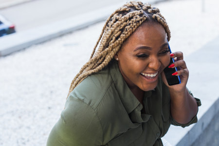Smiling african american woman with braids talking on smartphone outdoors. Concept of joy, friendship and positive communication.の写真素材