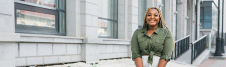 Confident african american woman standing against city architecture smiling. Concept of positivity, urban lifestyle and self-confidence. Banner with copy spaceの写真素材