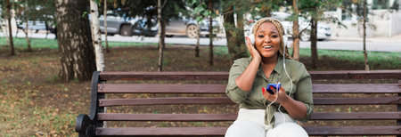 African american woman enjoying music with headphones and smiling at her phone on a bench. Joy, relaxation, and enjoying technology in everyday life. Banner with copy spaceの写真素材