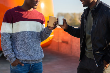 Two african american friends clink coffee cups on a sunny afternoon. Concept of friendship, connection, street lifestyle and casual communication.の写真素材