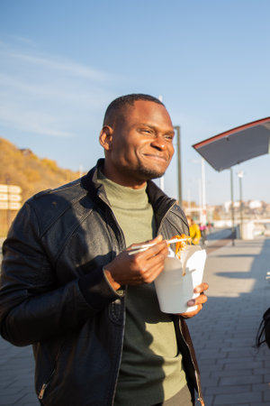 African american Man eating noodles from a takeaway box on the street. Concept of food culture,...