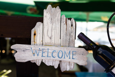 Rustic wooden welcome sign hanging near wine bottle. Hospitality greeting, vintage decor, and cozy outdoor restaurant charm.の写真素材