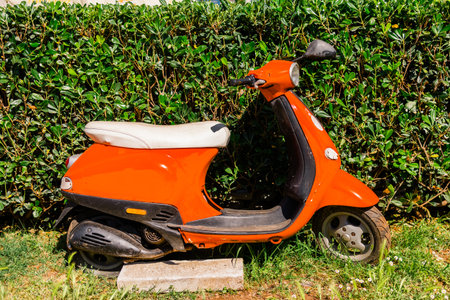Orange scooter parked beside green hedge under bright sunlight. Urban mobility, Mediterranean atmosphere, and retro travel aesthetic.の写真素材