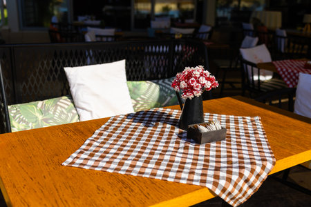 Rustic cafe table with checkered cloth and artificial flowers. Cozy outdoor setting with pillows and...