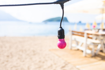 Pink light bulb hanging above beach sand. Decorative lighting element with blurred seaside background and wooden terrace.の写真素材